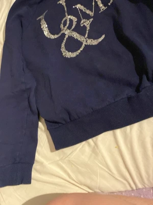 Mörkblå hoodie med paljetter - Mörkblå hoodie med glittrigt paljettmönster i silver på bröstet. Klassisk passform med ribbade muddar vid ärmslut och nederkant. Perfekt för dig som vill sticka ut lite extra med snygga detaljer.