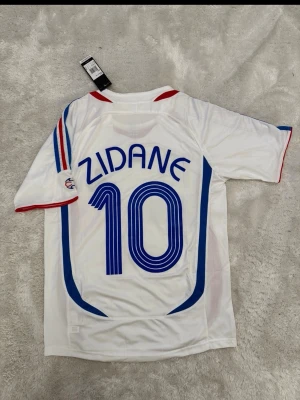 Frankrike Zidane matchtröja Adidas - Vit matchtröja från Adidas med blå och röda detaljer, nummer 10 och Zidane på ryggen. Tröjan har korta ärmar, broderad fransk landslagslogga och Adidas-logga på bröstet. Tillverkad i lätt och ventilerande material, perfekt för fotboll.