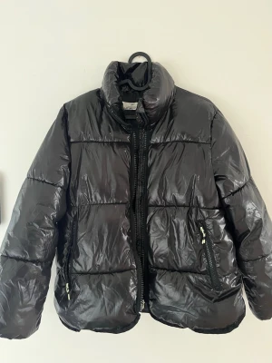 Svart glansig pufferjacka. Ungdom/barn - Säljer en svart, glansig pufferjacka med hög krage och dragkedja framtill. Jackan har två snedställda fickor med dragkedja och quiltad design. Perfekt för kalla dagar och ger en cool streetwear-look. Namnad på storlekslappen. 