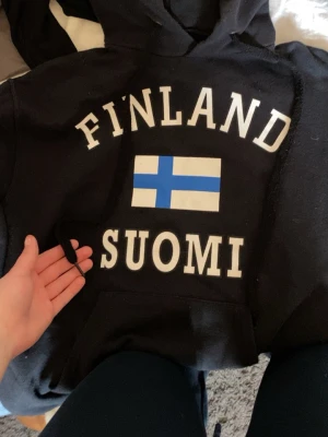 Svart Finland hoodie med flagga - Säljer en svart hoodie med texten 'FINLAND SUOMI' och en stor finsk flagga på bröstet. Klassisk huva, snören och magficka framtill. Perfekt för dig som vill visa kärlek till Finland och även kan användas på Finlands matcher!