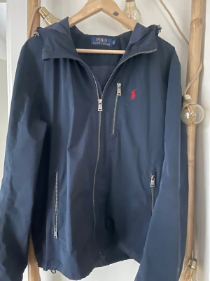 Marinblå vindjacka Polo Ralph Lauren - Snygg marinblå vindjacka från Polo Ralph Lauren med huva, dragkedja framtill och två fickor med dragkedja. Klassisk röd logga på bröstet. Perfekt för blåsiga dagar och enkel att matcha med olika outfits.