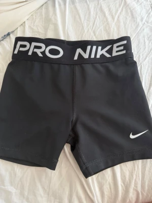 Svarta Nike Pro träningstights - Svarta korta träningstights från Nike Pro med bred resår i midjan där det står 'PRO NIKE' i vitt. Tightsen har en tight passform och är tillverkade i ett stretchigt funktionsmaterial som passar perfekt för träning. Är osäker på storlek men skulle ändå säga XS/XXS nypris 350kr