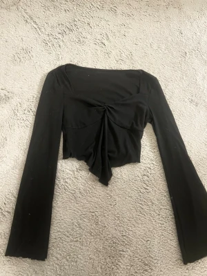 Svart croppad topp med mönster vid bysten - Trendig svart croppad topp med långa ärmar och snygg snörningsdetalj framtill. Toppen har en figurnära passform och är tillverkad i mjukt material som känns bekvämt mot huden. Perfekt för att lyfta vilken outfit som helst.