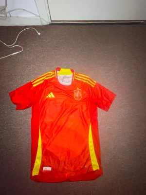 Spaniens matchtröja Adidas röd M - Säljer en röd matchtröja från Adidas för Spaniens landslag. Tröjan har gula detaljer på axlar och sidor, samt landslagets emblem på bröstet. Tillverkad i lätt och ventilerande material, perfekt för fotboll eller träning.