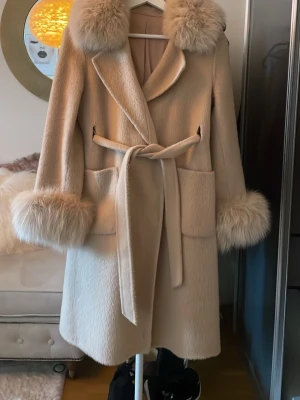 Beige kappa med äkta pälsdetaljer - Stilren beige kappa med stora fickor, knytskärp i midjan och fluffiga äkta pälsdetaljer vid krage och ärmslut. Kappan har en rak passform och är perfekt för kyliga dagar. Materialet ser ut att vara ullblandning och fuskpäls.