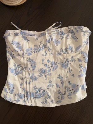 Blommig korsettopp med snörning - Korsettopp i vitt med blått blommönster och snörning upptill. Toppen är axelbandslös. 