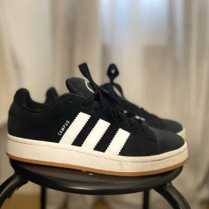 Adidas Campus svarta sneakers - Klassiska Adidas Campus sneakers i svart mocka med vita läderdetaljer och de ikoniska tre ränderna på sidan. Skorna har svart snörning, vit mellansula och gummifärgad yttersula. Perfekt för en stilren och sportig look.