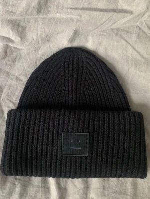 Svart ribbstickad mössa från Acne Studios - Säljer en svart ribbstickad mössa från Acne Studios med det klassiska ansiktslogotypen framtill. Mössan har bred uppvikt kant och är tillverkad i mjukt, tjockt material som håller värmen. Perfekt accessoar för kalla dagar.