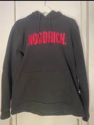 Svart HOODRICH hoodie med röd logga - Svart hoodie från HOODRICH med stor röd logga på bröstet. Klassisk känguruficka framtill, justerbar huva med snören och ribbade muddar. Tillverkad i mjukt sweatshirtmaterial som är perfekt för en avslappnad streetwear-look.