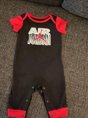 Air Jordan svart och röd sparkdräkt - Säljer en svart sparkdräkt från Air Jordan med röda ärmar och muddar. Tryck med Air Jordan-logga och den klassiska Jumpman-symbolen på bröstet. Kortärmad modell med tryckknappar i grenen för enkel påklädning. Perfekt för dig som gillar sportig stil.