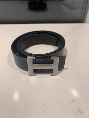 mörkblått skärp med H-spänne - Snyggt mörkblått skärp med ikoniskt H-format metallspänne i silver. Skärpet är gjort i slätt skinn och har vita kontrastsömmar längs kanterna. Perfekt accessoar för att lyfta din outfit.