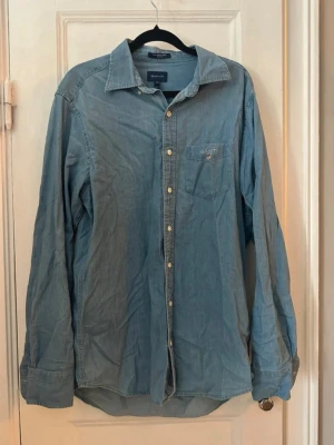 Blå skjorta från Gant  - Klassisk blå skjorta från Gant i mjuk bomull. Skjortan har lång ärm, bröstficka med liten logga och knappar framtill. Perfekt till jeans eller chinos för en avslappnad men snygg look.