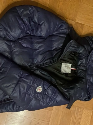 Mörkblå dunjacka från Moncler - Snygg mörkblå dunjacka från Moncler med klassisk quiltad design och hög krage. Jackan har dragkedja framtill, innerfoder med serietidningsmönster och Moncler-logga på bröstet. Perfekt för kalla vinterdagar och riktigt trendig stil.