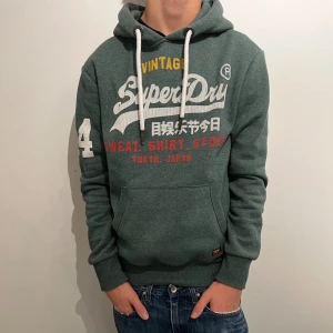 Grön Superdry hoodie med tryck - Säljer en grön hoodie från Superdry med stort vitt och rött tryck framtill, samt siffran 4 på ärmen. Tröjan har huva med vita snören och en klassisk känguruficka. Perfekt för dig som gillar streetwear och snygga detaljer.