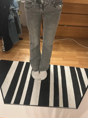 Grå bootcut jeans med kontrastsömmar - Säljer ett par grå bootcut jeans med snygga kontrastsömmar och klassiska fem fickor. Jeansen har en låg midja med två knappar.