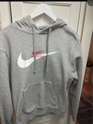 Grå Nike hoodie med logga - Snygg grå hoodie från Nike med stor vit och röd logga på bröstet och texten 'Nike Athletic Dept.' i rött. Klassisk känguruficka framtill, justerbar huva med dragsko och mjukt material. Perfekt för en sportig och avslappnad stil.