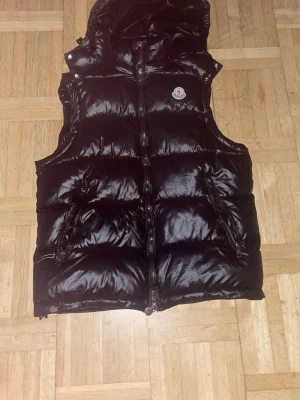 Svart dunväst från Moncler - Snygg svart dunväst från Moncler med glansig finish och huva. Västen har två dragkedjefickor fram och klassisk Moncler-logga på bröstet. Perfekt för lager-på-lager och håller dig varm under kyliga dagar. Stängs med dragkedja framtill.