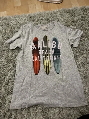 Grå t-shirt med surfbrädor och text - Snygg grå t-shirt från Lindex med tryck av tre surfbrädor i rött, gult och blått samt texten 'Malibu Beach California' över bröstet. T-shirten har rund hals och korta ärmar, perfekt för en avslappnad stil.