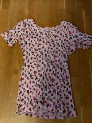 Blommig ribbad topp med knappar - Söt ljusrosa ribbad topp med småblommigt mönster i rött, blått och gult. Toppen har korta ärmar och dekorativa knappar framtill. Perfekt för dig som gillar en romantisk och avslappnad stil.