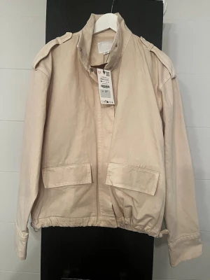 Zara Beige jacka i storlek XS - Stilren beige vindjacka från Zara med hög krage, axelklaffar och stora fickor framtill. Jackan har dragsko i nederkant och tryckknappar vid halsen. Perfekt för dig som vill ha en enkel men trendig look. Materialet är bomullsmix.
