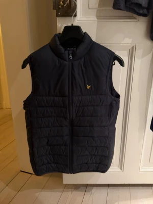 Svart dunväst från Lyle & Scott - Snygg svart dunväst från Lyle & Scott med gul logga på bröstet. Västen har hög krage, dragkedja framtill och quiltad design. Perfekt att slänga över en hoodie eller tröja för extra värme. Klassisk och stilren look.