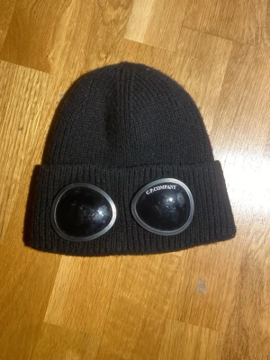 Svart mössa med goggles C.P. Company - Säljer en svart ribbstickad mössa från C.P. Company med ikoniska goggles framtill. Mössan är tillverkad i mjukt material och har en uppvikt kant. Perfekt för dig som vill sticka ut med en streetwear-inspirerad look.