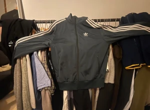 Smaragdgrön/Mörkgrön Adidas track jacket - Snygg smaragdgrön track jacket från Adidas med klassiska vita ränder längs ärmarna och dragkedja framtill. Jackan har ståkrage, två fickor med dragkedja och Adidas logga på bröstet. Priset är inte satt i sten!