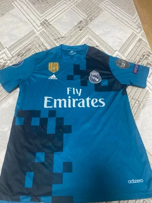 Real Madrid bortatröja Adidas blå - Säljer en blå Real Madrid bortatröja från Adidas med pixelmönster i svart, Champions League och FIFA-märken på ärmarna samt klubbmärke och Fly Emirates-tryck på bröstet. Tillverkad i lätt och ventilerande material, perfekt för fotboll eller träning. Ronaldo tröja, passar mest stl M tror jag.