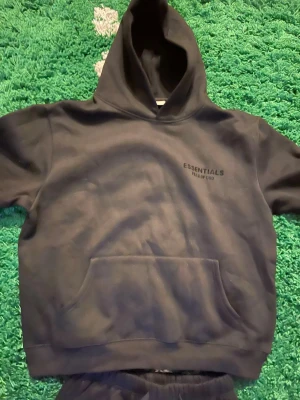 Svart Essentials Fear of God hoodie och byxor - Säljer en svart hoodie från Essentials Fear of God med diskret logga på bröstet. Hoodien har stor huva och en klassisk magficka. Tillverkad i mjukt material som känns skönt mot huden. Perfekt för dig som gillar streetwear och enkel stil.