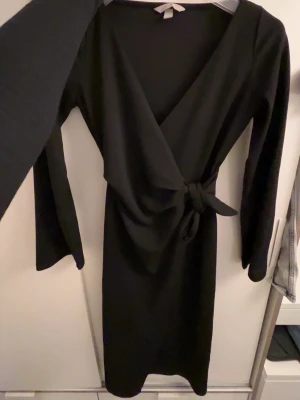 Svart omlottklänning med knyt - Stilren svart omlottklänning med lång ärm och knyt i midjan. Klänningen har v-ringad hals och en elegant, figurnära passform. Perfekt för dig som gillar en klassisk look med en twist. Materialet känns mjukt och följsamt. Kläningen är köpt på hm och endast använd 1 gång 
