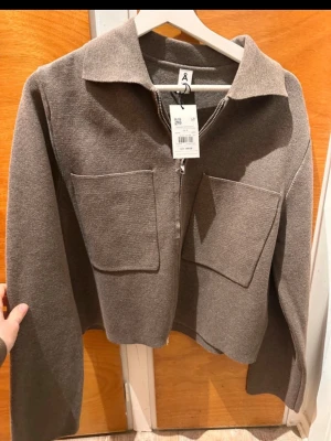 Brun zip cardigan med stora fickor - Snygg brun cardiganjacka från åhlens med dragkedja framtill och två stora fyrkantiga fickor på bröstet. Jackan har en rak, boxig passform och klassisk krage. Materialet är stickat och ger en mjuk känsla. Perfekt för lager på lager och stilrena outfits.