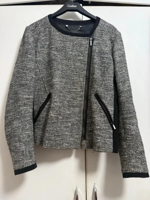 Gråmelerad kavaj med dragkedja - Stilren gråmelerad kavaj med asymmetrisk dragkedja och svarta detaljer vid krage, fickor och ärmslut. Rak passform och rund halsringning. Materialet är vävt tyg som ger en modern look. 