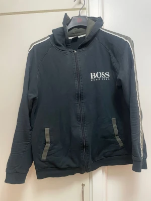 Svart hoodie från Hugo Boss - Snygg svart hoodie med dragkedja från Hugo Boss. Tryckt logga på bröstet, grå detaljer på fickorna och vita ränder längs ärmarna. Perfekt för en avslappnad och trendig stil. Mjuk bomullsblandning och huva för extra komfort.