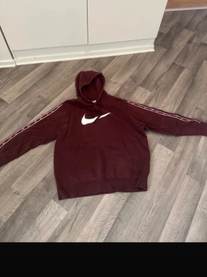 Vinröd Nike hoodie med logga - Säljer en vinröd hoodie från Nike med stor vit logga på bröstet och snygga ränder längs ärmarna. Hoodien har huva och en stor magficka framtill. Perfekt för dig som gillar sportig och avslappnad stil.