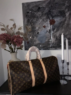 Louis Vuitton Keepall Monogram väska - Stilren och rymlig Louis Vuitton Keepall handväska i klassiskt brunt monogramcanvas med beige läderdetaljer och guldfärgade metalldelar. Väskan har dubbla handtag, dragkedja och innerfack med dragkedja. Perfekt för dig som gillar lyxig och tidlös design.