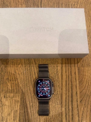 Apple Watch Ultra med svart milanese-armband - Apple Watch Ultra med fyrkantig svart boett och svart milanese-armband i rostfritt stål. Smartklockan har en stor och tydlig skärm med digital urtavla och levereras i originalförpackning. Perfekt för dig som vill ha stilren teknik på handleden.