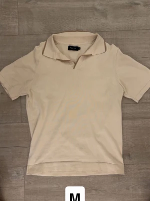 Beige piké t-shirt från Riley - Säljer en stilren beige piké t-shirt från Riley. T-shirten har korta ärmar, klassisk krage och är tillverkad i mjuk bomull. Perfekt för en clean och avslappnad look.