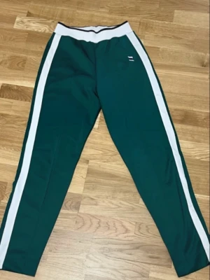 Gröna mjukisbyxor från Zara - Mörkgröna mjukisbyxor från Zara med bred vit resår i midjan och vita sidoränder längs benen. Sportig look med avslappnad passform, perfekta för chill eller träning. Tillverkade i mjukt material för extra komfort.