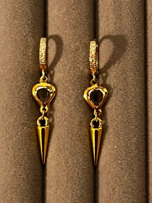 Guldpläterade örhängen med hjärta och spets - Snygga guldpläterade örhängen med gnistrande stenar på bygeln, hjärtformad svart sten och hängande spetsig detalj. Örhängena har en modern och edgy design som sticker ut. Perfekta för dig som vill ha något unikt och trendigt i smyckeskrinet.