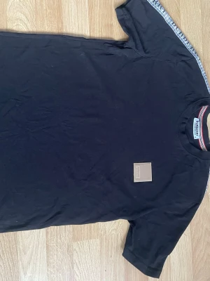 Svart Burberry t-shirt med logga - Snygg svart t-shirt från Burberry med klassisk logga på bröstet och randiga detaljer i halsen. T-shirten har korta ärmar och en stilren design med diskret Burberry-märke. Perfekt för dig som gillar exklusiva streetwear-plagg.