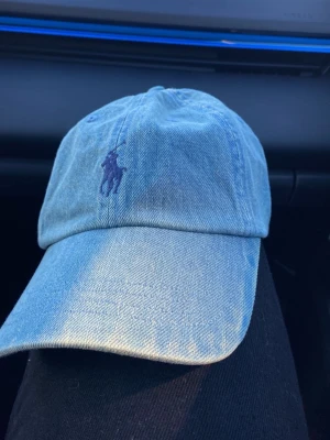 Ljusblå denimkeps från Polo Ralph Lauren - Säljer en ljusblå keps i denim från Polo Ralph Lauren med klassisk böjd skärm och broderad logga i mörkblått framtill. Kepsen har justerbar rem bak och är tillverkad i bomull. Perfekt accessoar för en avslappnad och trendig look.