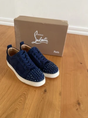 Blåa Louboutin sneakers med nitar - Snygga mörkblå sneakers från Christian Louboutin i mocka med coola nitar på tån och klassisk röd sula. Skorna har vita sulor och blå snören, samt en låg profil som ger en clean look. Perfekta för dig som vill sticka ut med exklusiva detaljer. Storlek 43 men passar 42. Priset är inte hugget i sten!  