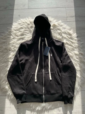 Svart hoodie från Polo Ralph Lauren - Svart hoodie med dragkedja från Polo Ralph Lauren. Tröjan har vit logga broderad på bröstet, vita snören i huvan och två fickor framtill. Perfekt för en avslappnad och stilren look.