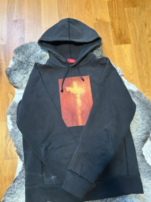 Svart Supreme hoodie med tryck - Supreme hoodie i svart med stor huva och dragsko. På bröstet finns ett rektangulärt tryck i rödorange toner som ger en unik vibe. Klassisk känguruficka framtill och ribbade muddar. Perfekt för dig som vill sticka ut med streetwear från Supreme.