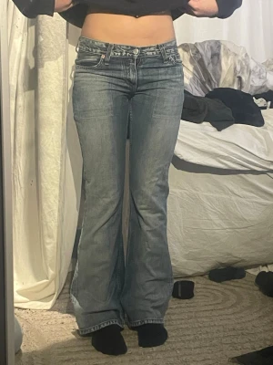 Blå bootcut jeans från Levi's - Säljer ett par klassiska blå bootcut jeans från Levi's med slitningar och låg midja. Jeansen har fem fickor, dragkedja och knapp framtill samt snygga detaljer på bakfickorna. Perfekta för en avslappnad och trendig look.