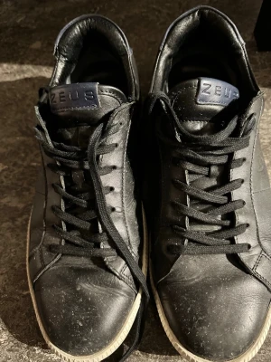 Svarta Zeus sneakers i läder - Svarta sneakers från Zeus med klassisk låg profil och svarta skosnören. Ovandelen är i slätt läder och sulan är vit med diskret mönster. Märkeslogga på plösen. Perfekta för dig som gillar stilrena och tidlösa skor.