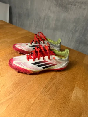 Adidas F50 - Säljer ett par snygga Adidas fotbollsskor med vit bas, röda detaljer och svarta ränder på sidan. Skorna har röda snören och insidan är limegrön. Yttermaterialet är syntetiskt och sulan är anpassad för spel på gräs. Perfekta för dig som vill sticka ut på planen! Använts ett par gånger så dem är lite smutsiga men inga problem annars. Dobbarna är som nya!