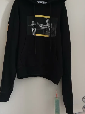 Svart hoodie med tryck från Off-White - Snygg svart hoodie från Off-White med grafiskt tryck framtill och gula diagonala ränder längs ärmarna. Klassisk huva med dragsko och känguruficka. Materialet känns mjukt och tjockt, perfekt för en streetwear-look. Hör av er vid frågor 