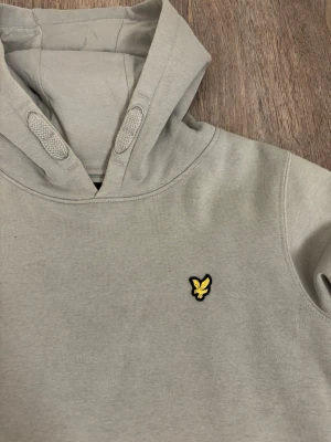 Ljusgrön hoodie från Lyle & Scott - Snygg ljusgrön hoodie från Lyle & Scott med klassisk gul örnlogga på bröstet. Hoodien har huva med dragsko och en stor känguruficka framtill. Tillverkad i mjukt bomullsmaterial som är skönt att ha på sig. Perfekt för en avslappnad och stilren look.