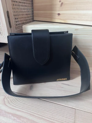 Svart axelväska från Jacquemus - Le bambino 24. Snygg svart axelväska från Jacquemus i stilren men annorlunda fyrkantig modell. Kvitto från inköp på hemsidan finns. Även dustbag finns. Fint skick!                                                                                    Width: 26,5 cm Height: 22 cm Depth: 9,5 cm Strap: 62 cm